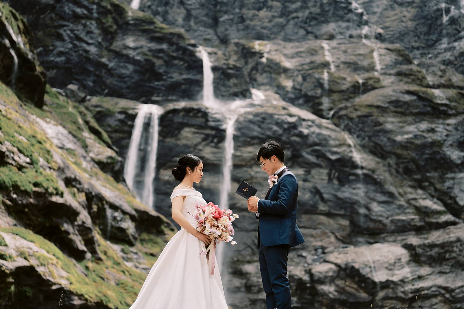 Queenstown New Zealand Heli Wedding Elopement Photographer クイーンズタウン　ニュージーランド　エロープメント 結婚式 | A couple exchanging vows in front of a waterfall, captured by a Queenstown Wedding Photographer, creating a scenic and intimate wedding moment.